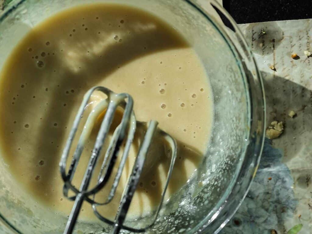 A glass bowl containing a creamy batter with air bubbles is shown. Two metal beaters are partially submerged in the mixture, resting against the side of the bowl. A textured, slightly messy surface is visible beside the bowl.